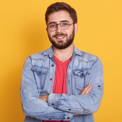 Indoor picture of cheerful handsome young man having folded hands, looking directly at camera, smiling sincerely, wearing casual clothes, posing isolated over yellow background. Emotions concept.